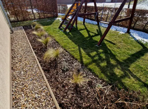 "Backyard garden featuring a mulched flower bed with ornamental grasses, a pebble pathway, and a wooden play structure casting a shadow on the lawn."