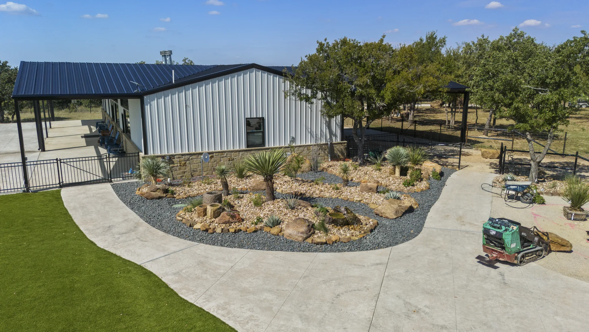 Aerial view of a boulder and agave bed in decomposed granite beside a concrete drive and barndominium