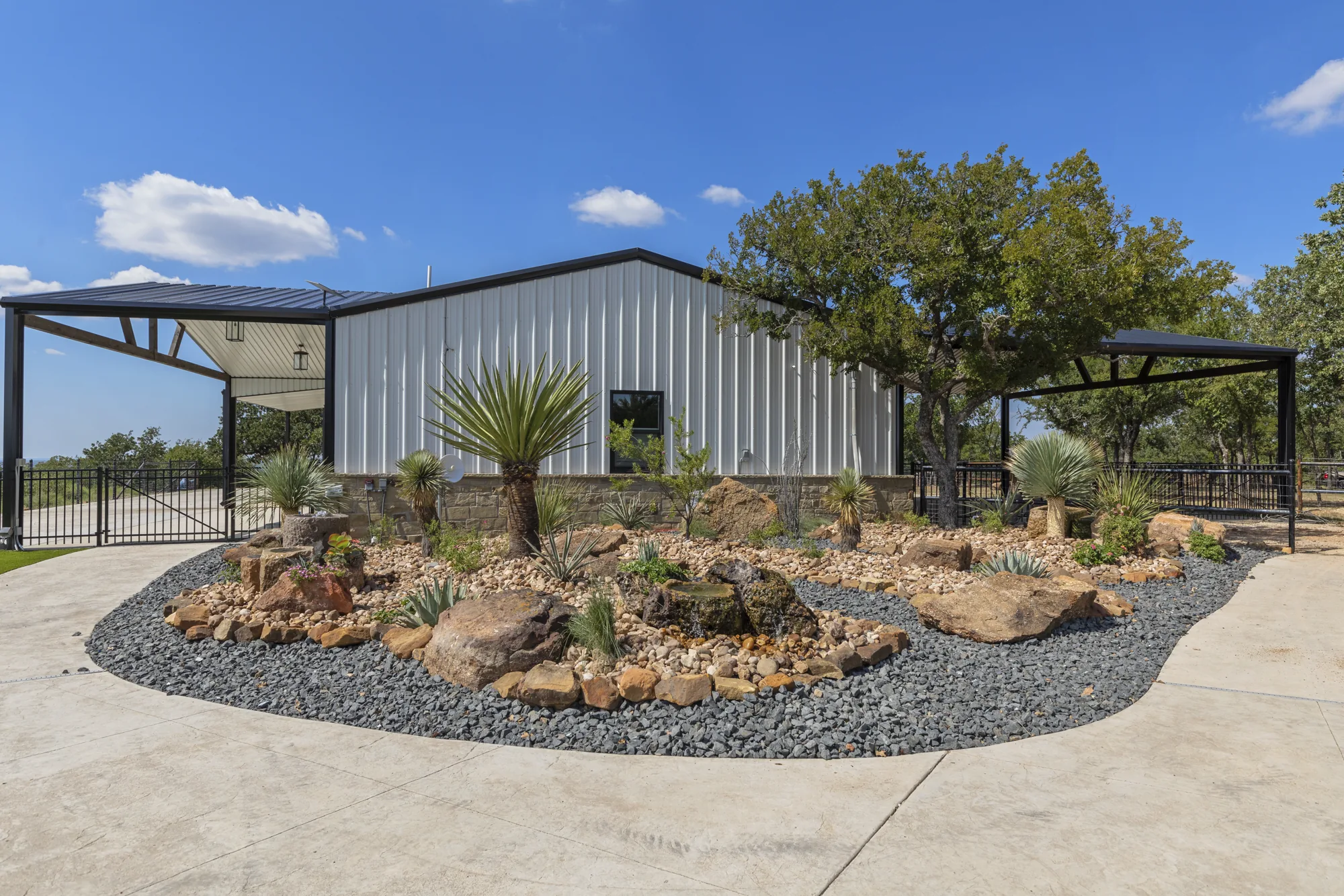 A boulder and agave island bed in decomposed granite with black rock mulch, in front of a white metal barndominium