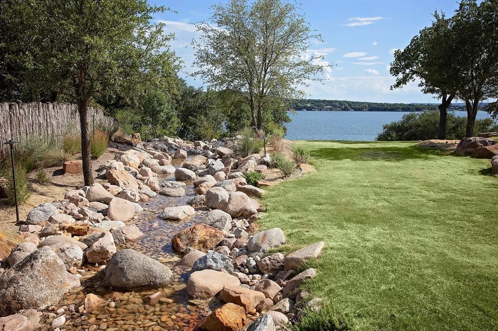 Boulder-lined dry creek bed running through a lakefront lawn to open water, with the far shore beyond