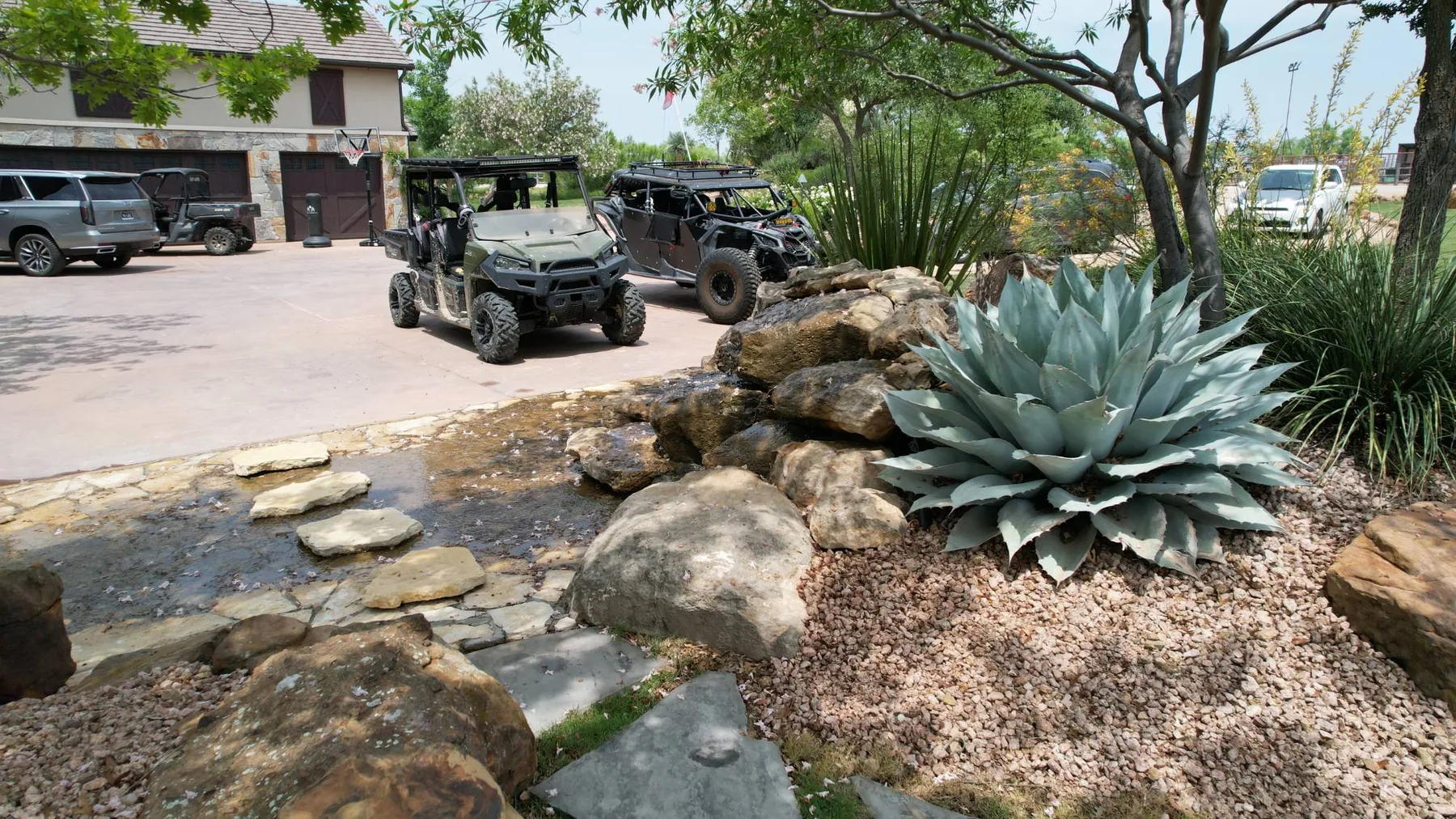 A boulder water feature crossed by flagstone stepping stones, with a large agave alongside and a stone barn beyond