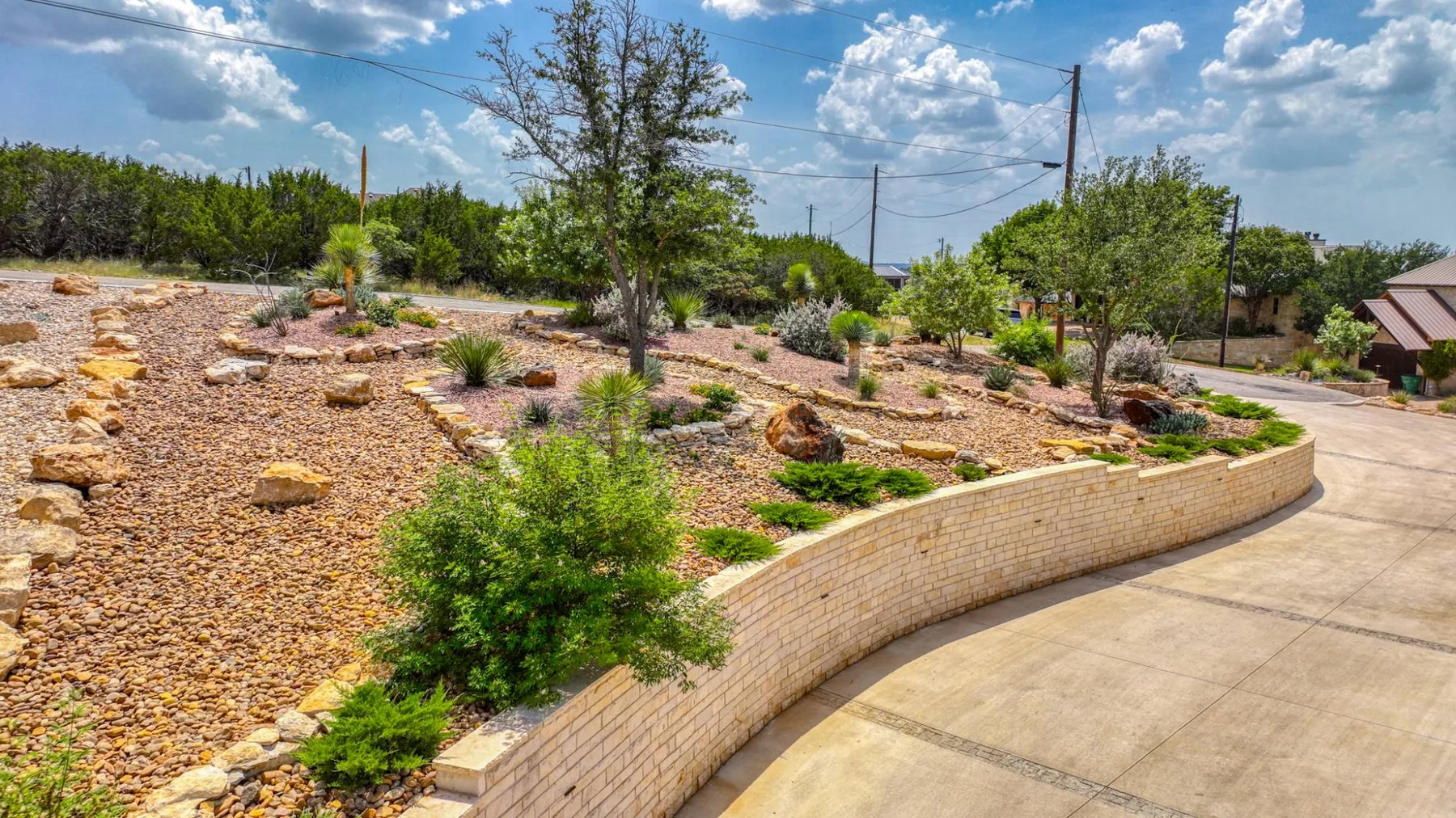 A long curved limestone retaining wall holding a broad gravel xeriscape bed planted with yucca, sotol and juniper