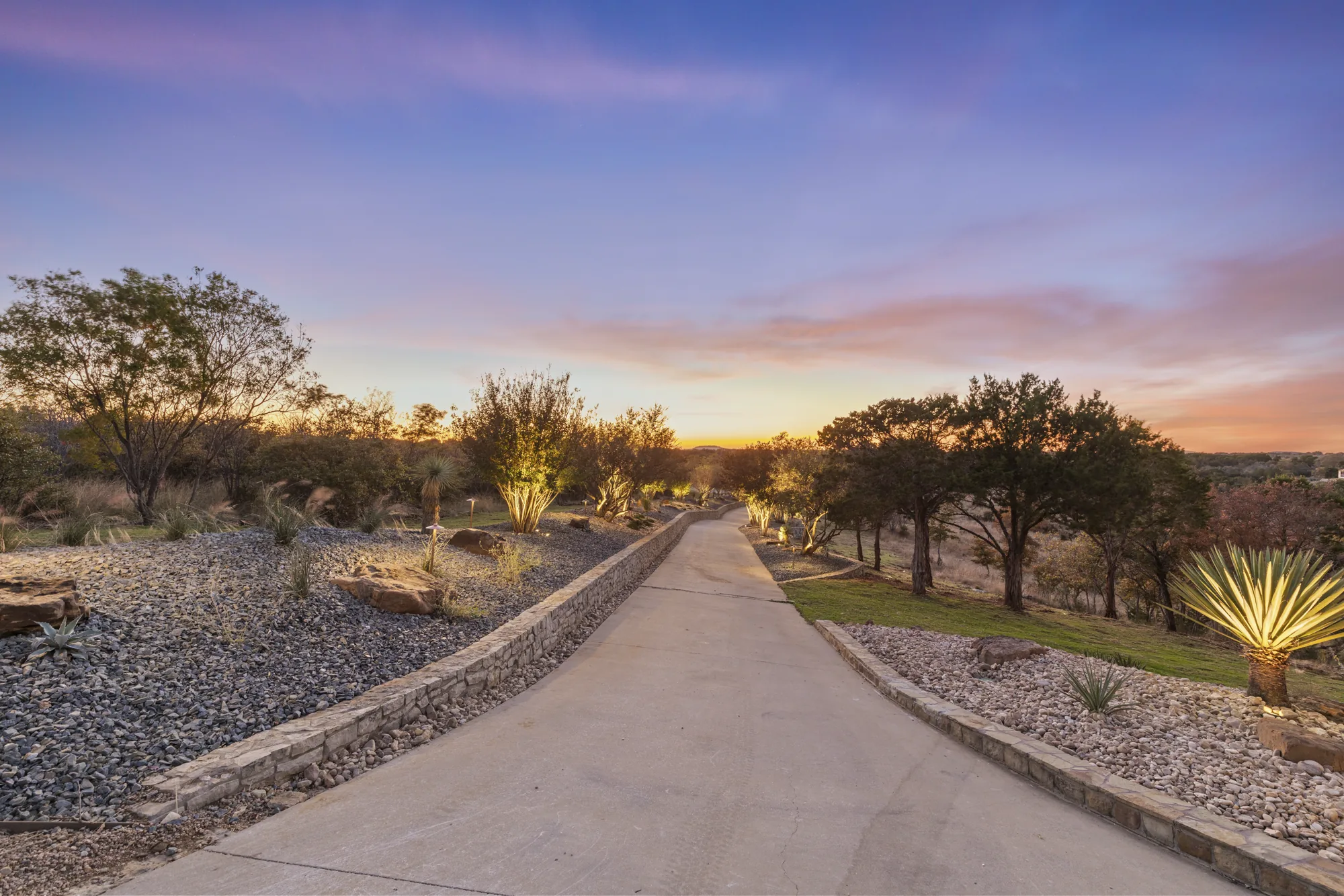 A long concrete drive at dusk between a stone retaining wall and rock mulch beds with young plantings