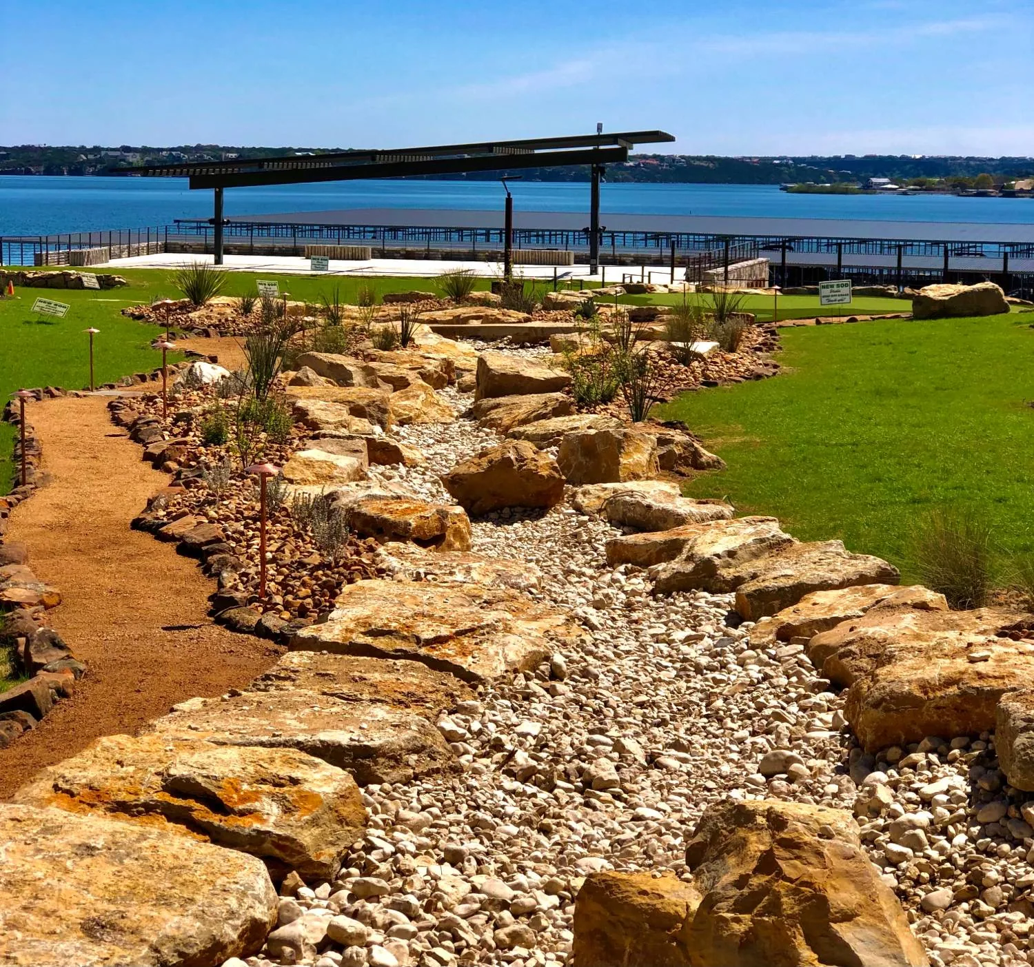 A wide boulder and cobble dry creek channel running down a lawn toward the lake, edged with copper path lights
