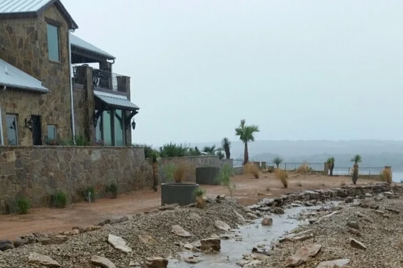 A stone-lined dry creek carrying running water past a lake house during rain, the lake grey behind it