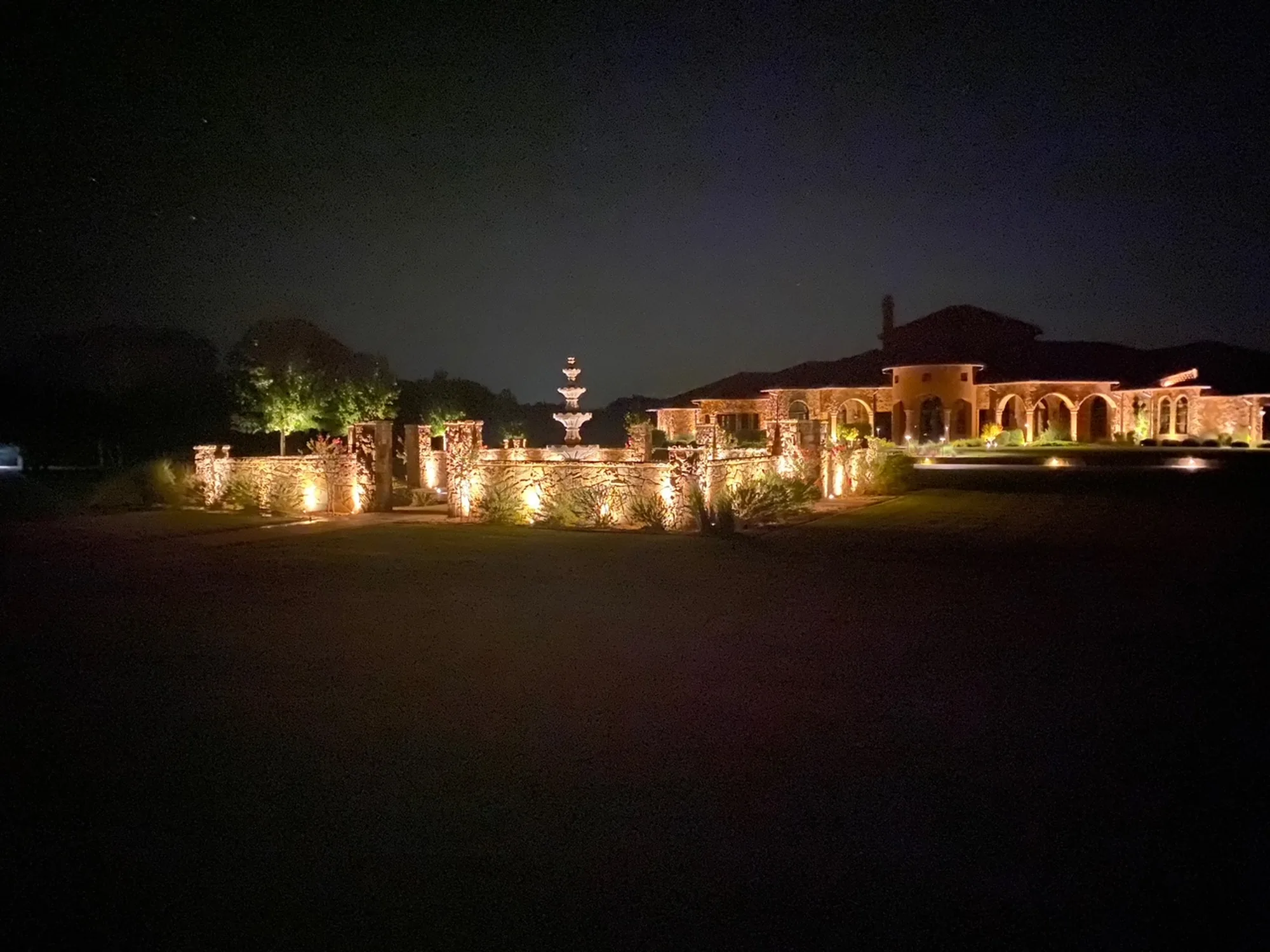 Stone entrance wall and columns washed in low-voltage light at night, with a tiered fountain and lit residence beyond