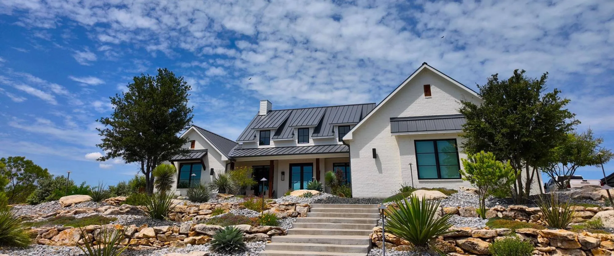 White brick farmhouse above dry-stacked limestone terracing, river-rock beds and yucca, with steps up to the entry