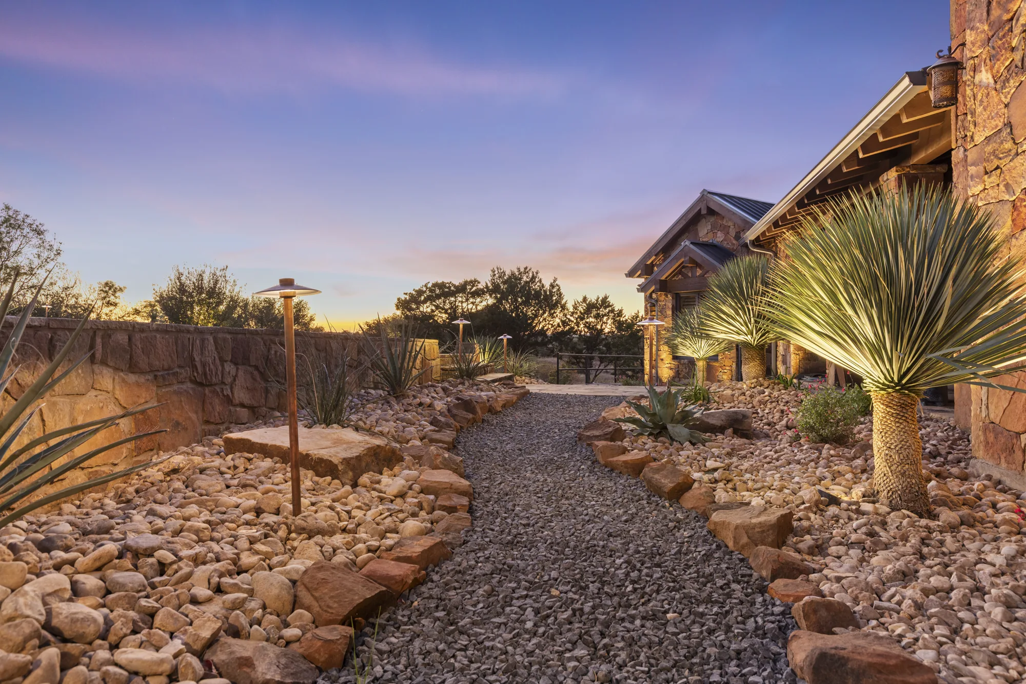A flagstone path between rock mulch beds at twilight, with an uplit yucca and a copper path light
