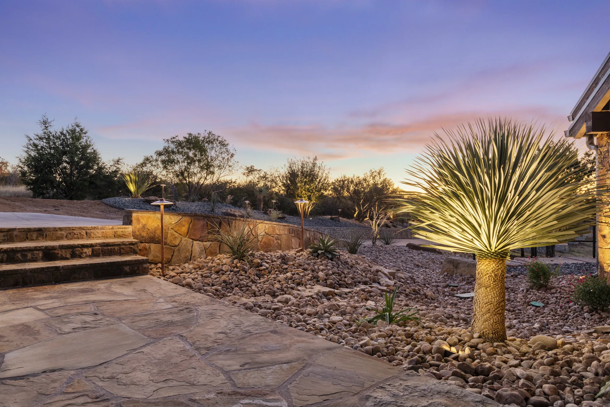 A flagstone patio and stone steps beside a stone retaining wall at twilight, with an uplit yucca