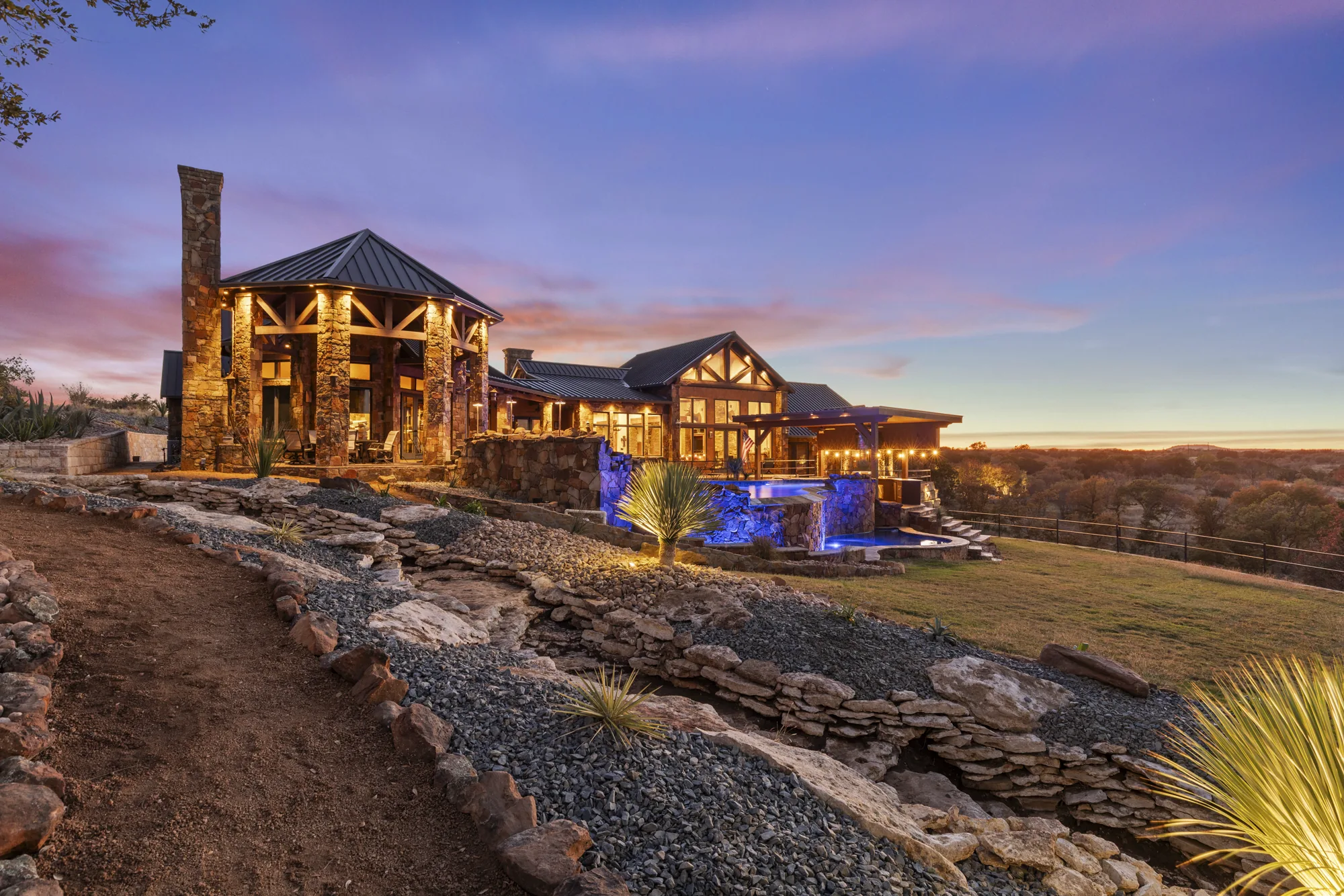 A lit ranch home and pool above dry-stacked stone terracing and boulder work on a hillside at twilight