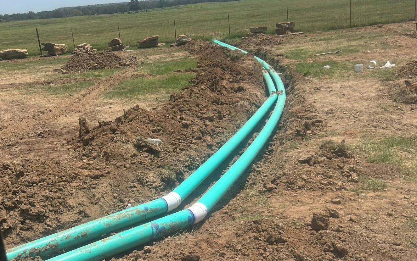 Twin teal irrigation mainlines laid in an open trench curving across pasture, spoil banked along the run