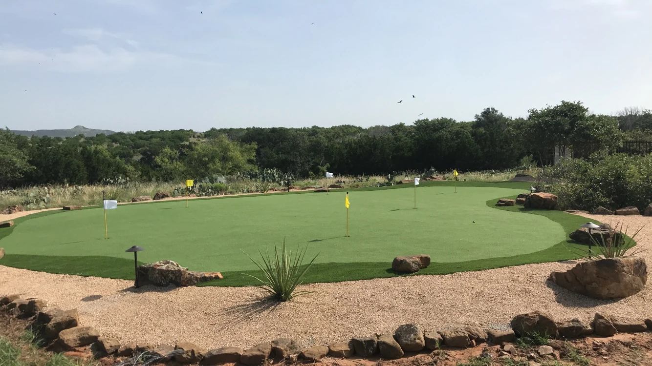 A synthetic putting green with five flags set into decomposed granite and boulders, with native brush and prickly pear behind