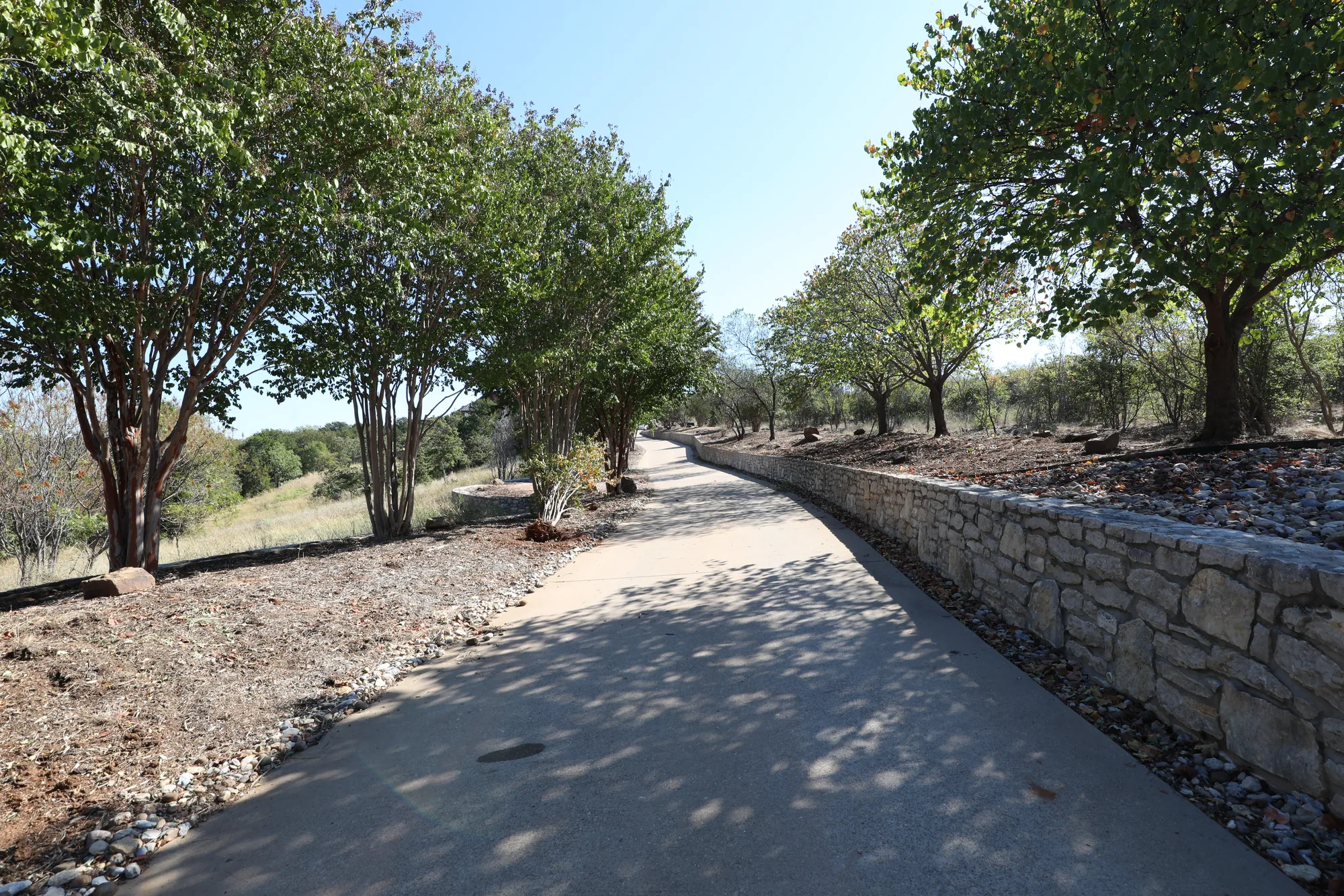 A paved drive curving past a dry-stacked stone retaining wall on a ranch property, trees lining the route