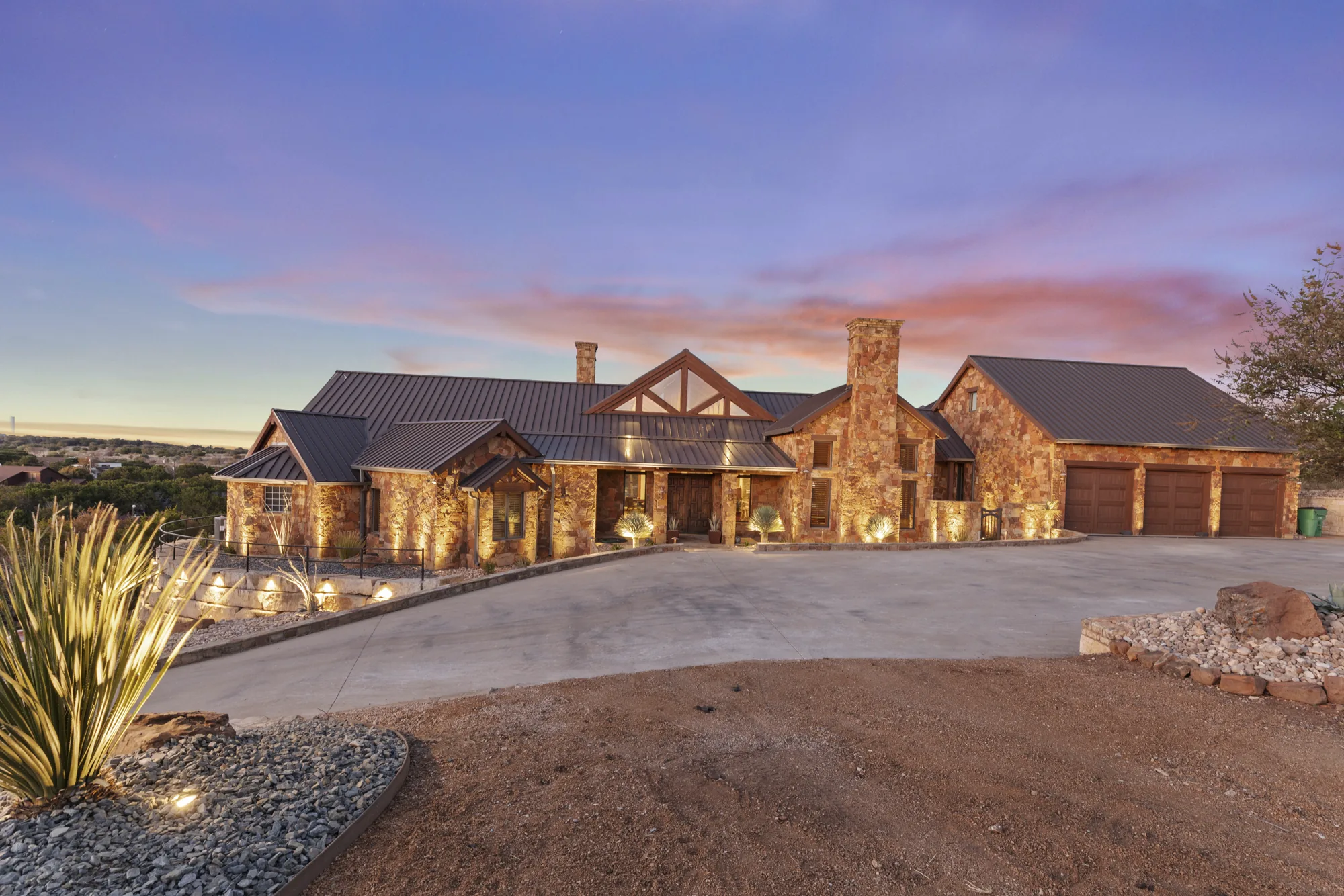 A stone ranch home lit at twilight above a circular concrete drive, with uplit agave in the foreground