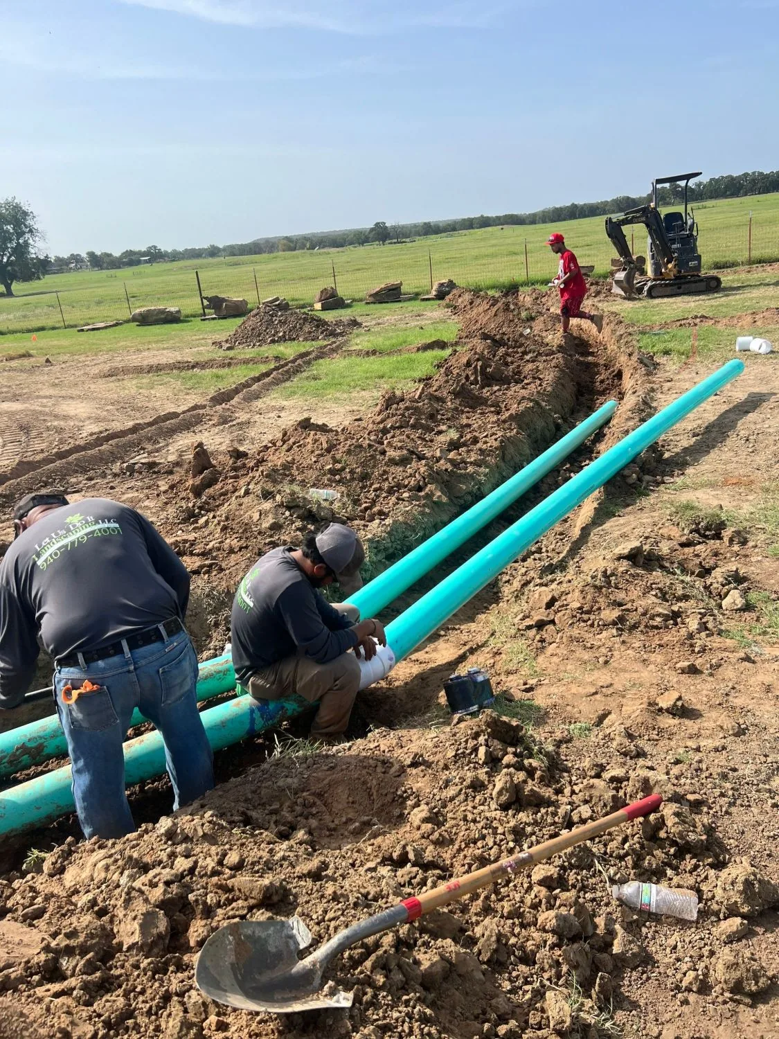 Two Let Us Do It crew members solvent-welding a coupling onto an irrigation mainline beside an open trench
