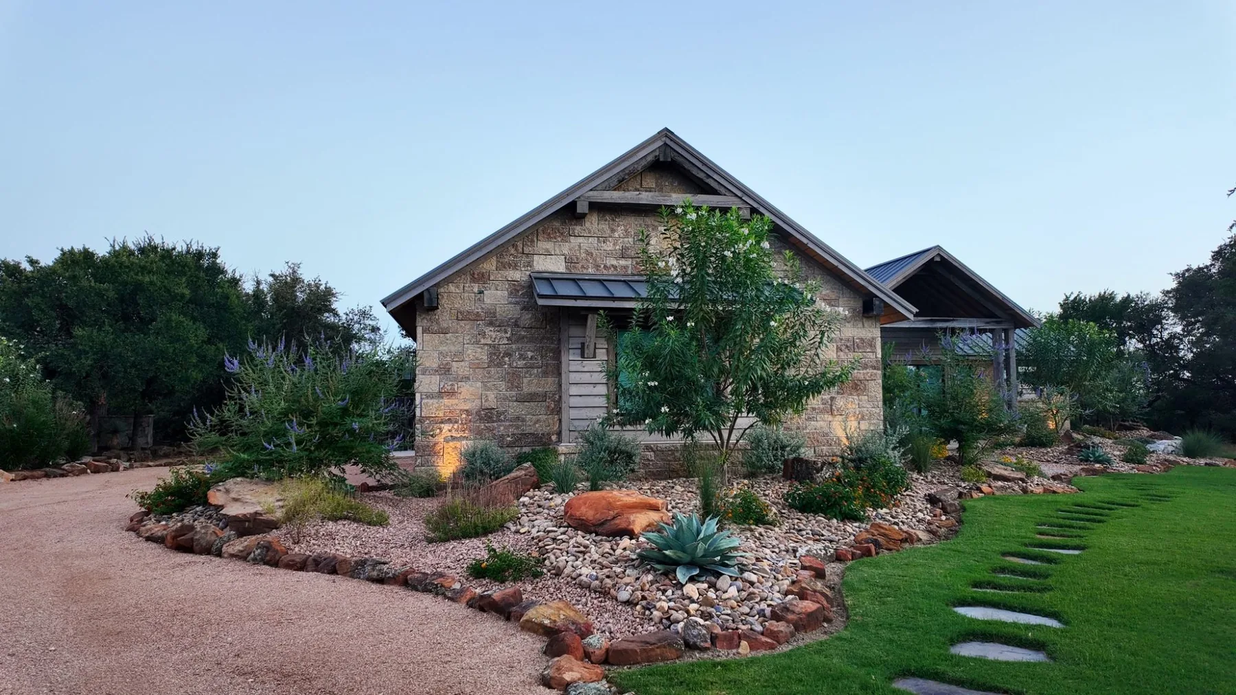 Stone home at dusk behind river-rock beds with agave and flowering vitex, a gravel drive and a stepping-stone lawn